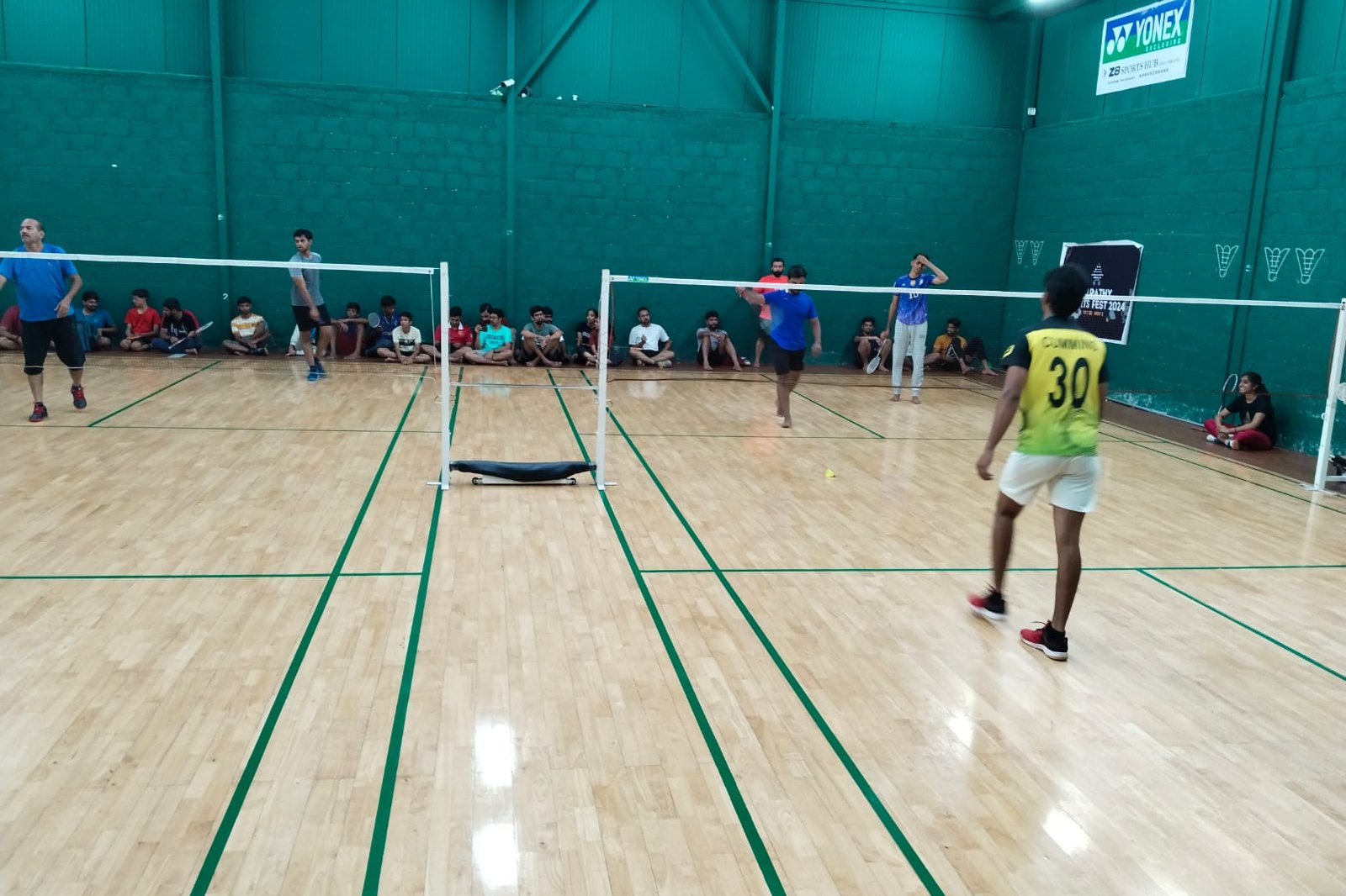 Players on an indoor wooden badminton court with spectators seated along the wall