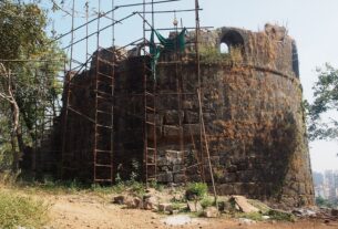 A bastion of Belapur Fort covered in scaffolding with Navi Mumbai buildings behind it