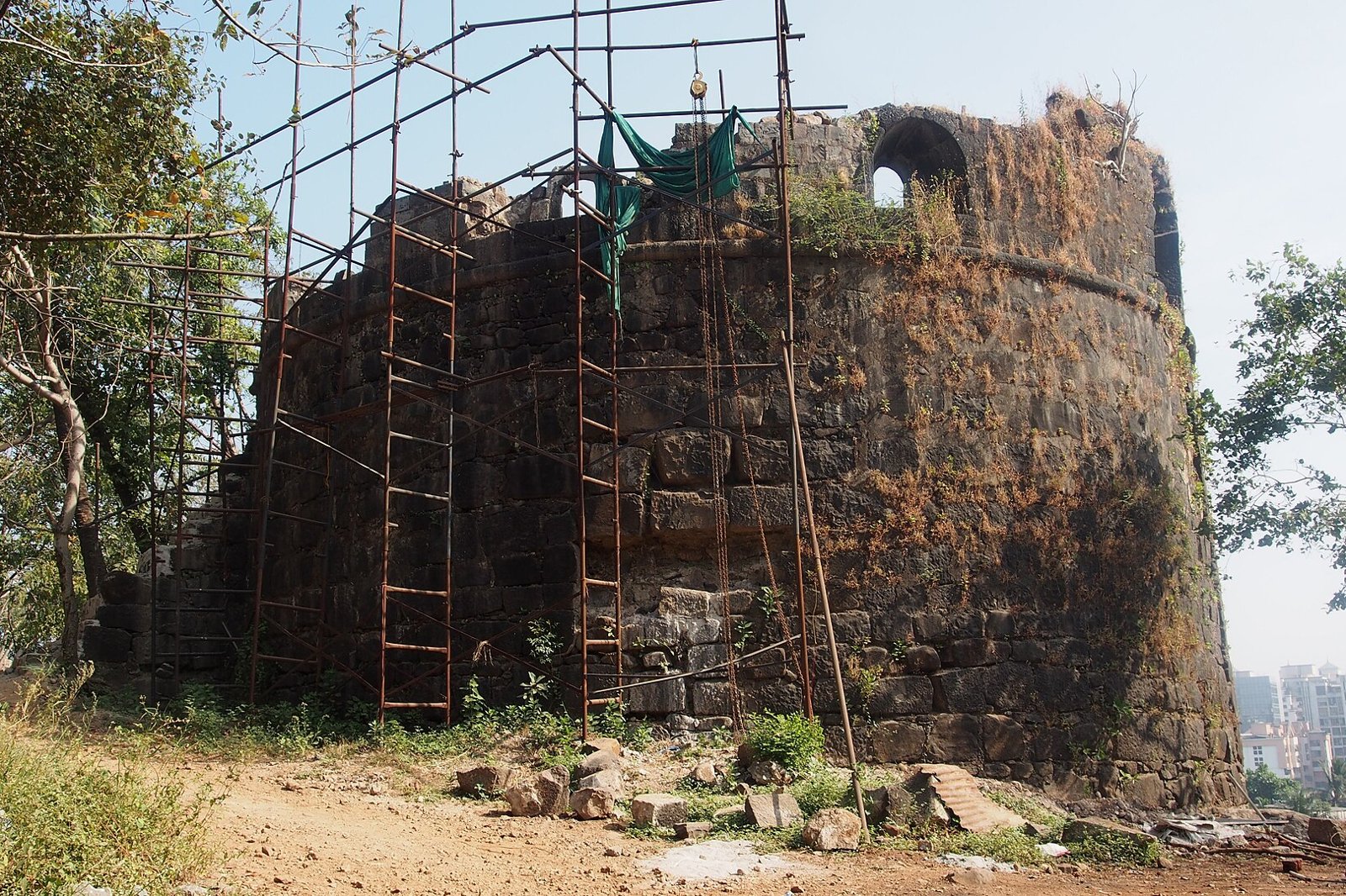 A bastion of Belapur Fort covered in scaffolding with Navi Mumbai buildings behind it