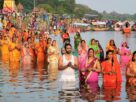 Women and men in bright clothes standing in knee deep water with hands folded during a Chhath Puja gathering