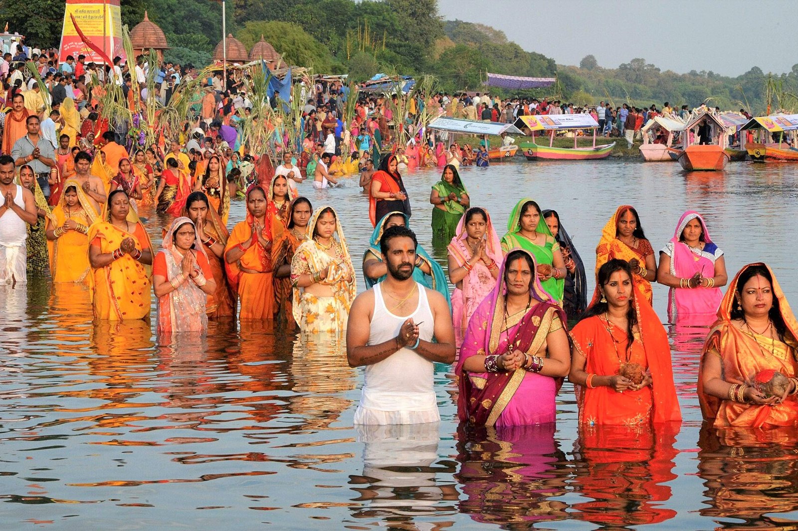 Women and men in bright clothes standing in knee deep water with hands folded during a Chhath Puja gathering