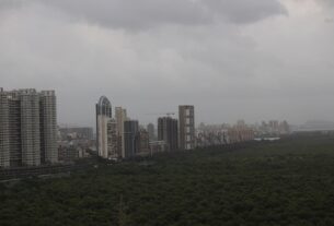 Cluster of tall apartment towers rising above dark green mangroves under a cloudy sky along Palm Beach Road in Navi Mumbai