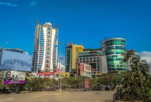 Tall apartment tower beside glass fronted commercial buildings and billboards under a clear blue sky in Vashi