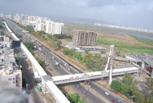 Aerial view of a busy road with a cable stayed footbridge, a partly built apartment tower and open land beyond in the Kharghar area