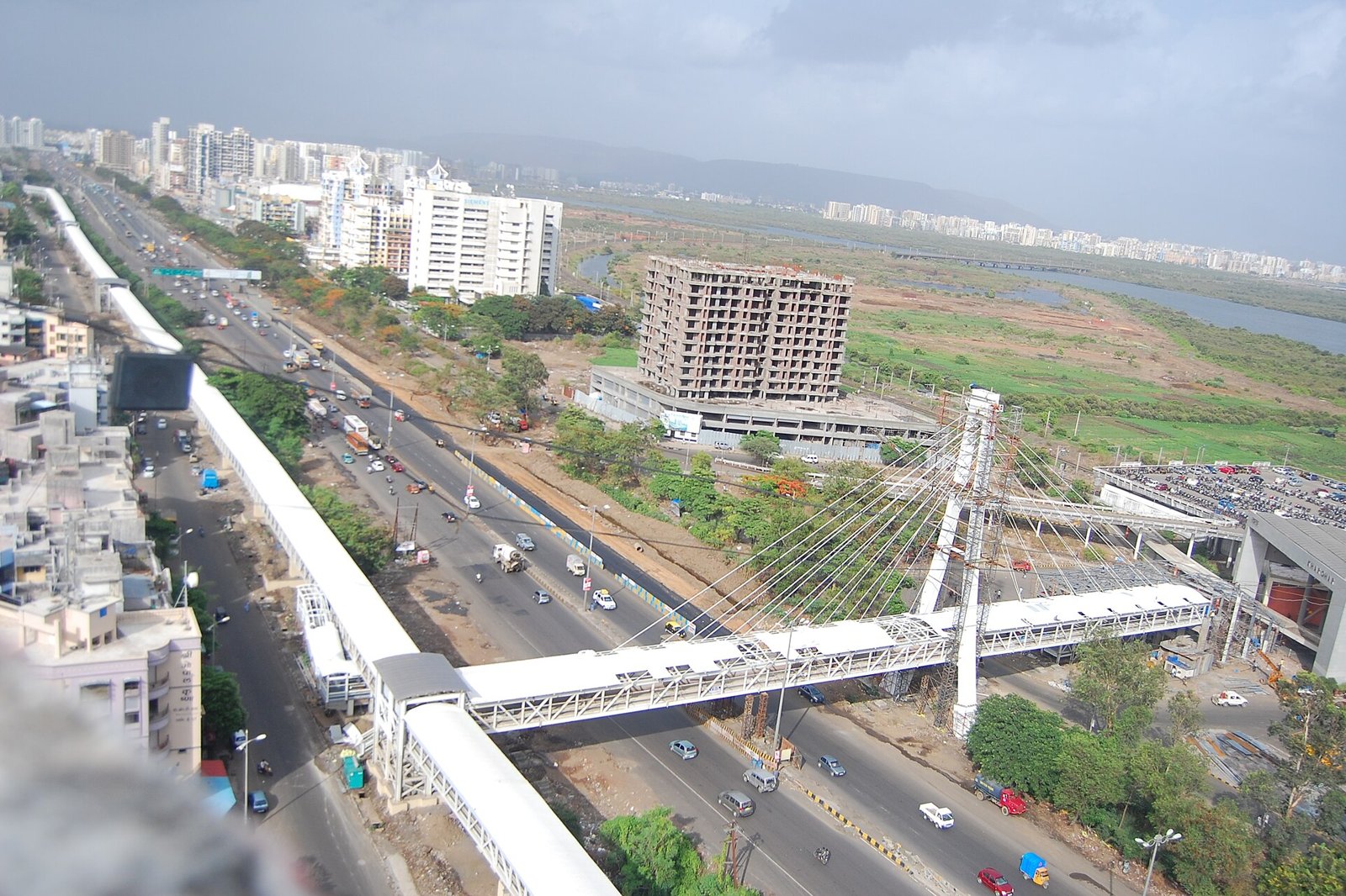 Aerial view of a busy road with a cable stayed footbridge, a partly built apartment tower and open land beyond in the Kharghar area
