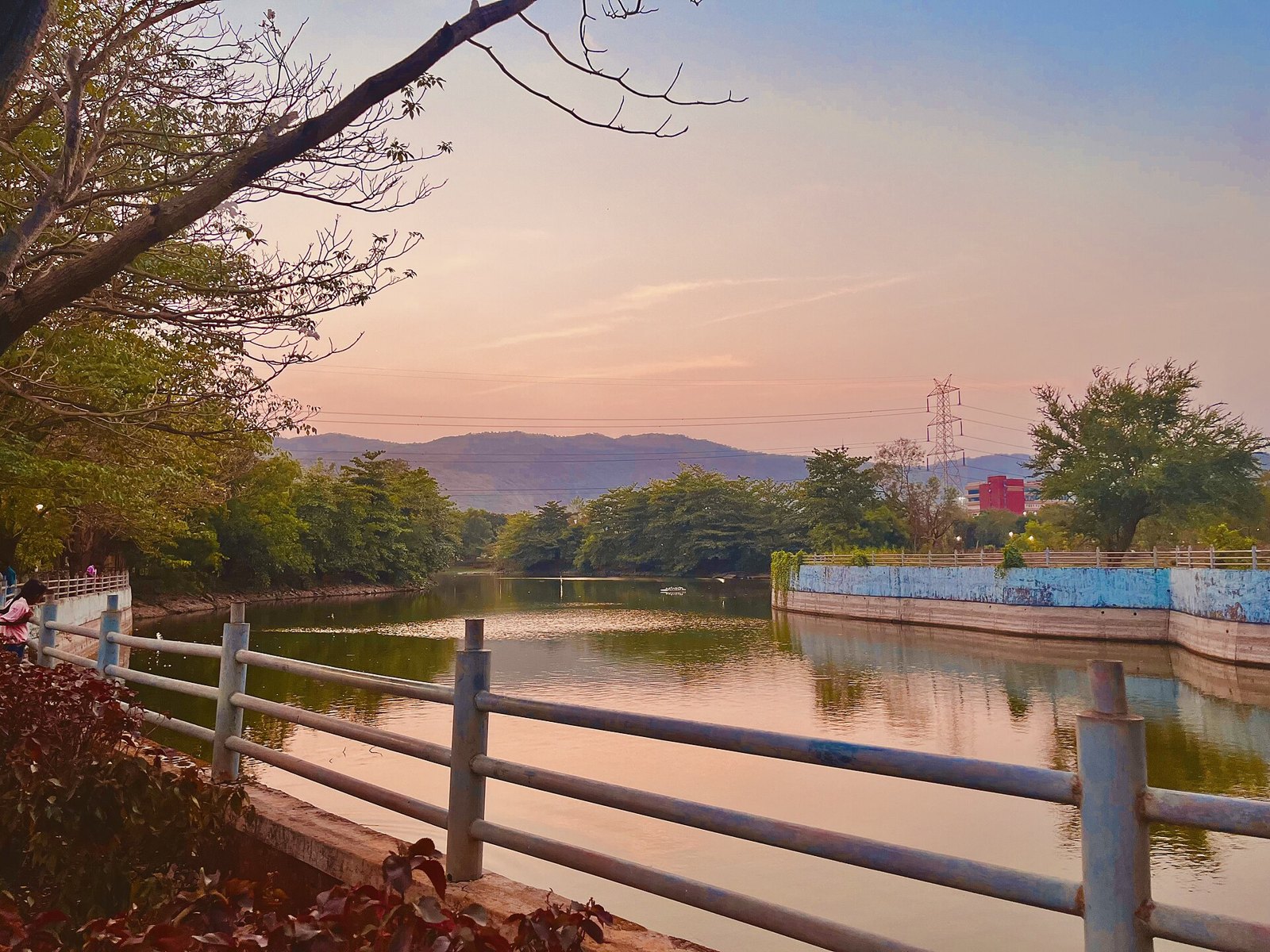 Artificial lake inside Central Park in Kharghar at dusk, with trees, a railing and hills in the background