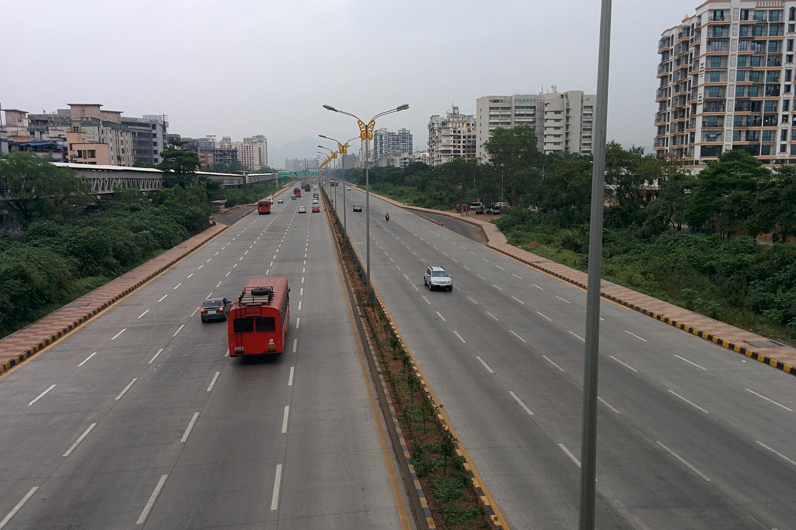 A wide multi lane highway with buses and cars passing apartment buildings and greenery along the Sion Panvel highway