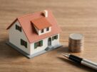 A small model house beside a stack of coins and a pen on a wooden desk