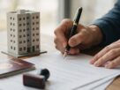A person signing a property document with a fountain pen beside a passport, a rubber stamp and a model apartment building