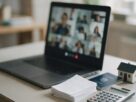 A laptop on a video call beside bank papers, a passport, a calculator and a small model house