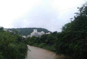 White apartment buildings on a wooded hillside beside a brown stream in Kharghar