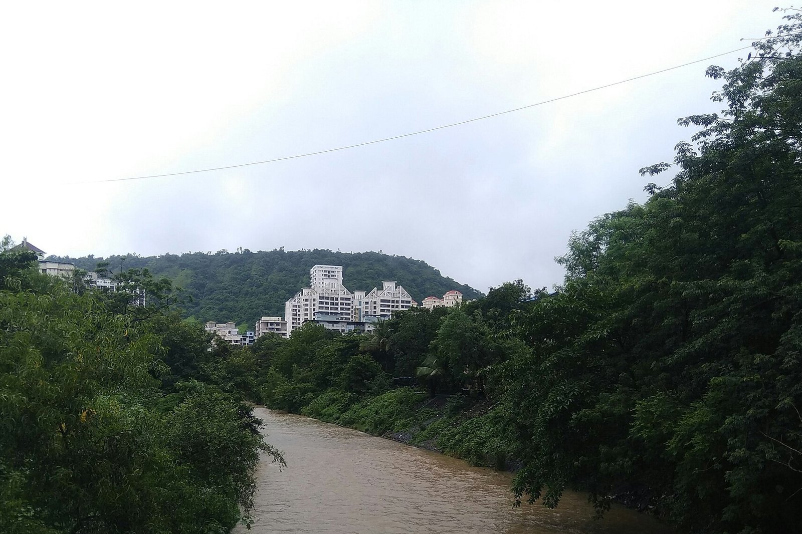 White apartment buildings on a wooded hillside beside a brown stream in Kharghar