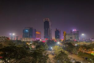 Tall buildings lit up at night in Vashi, seen over trees and a car park