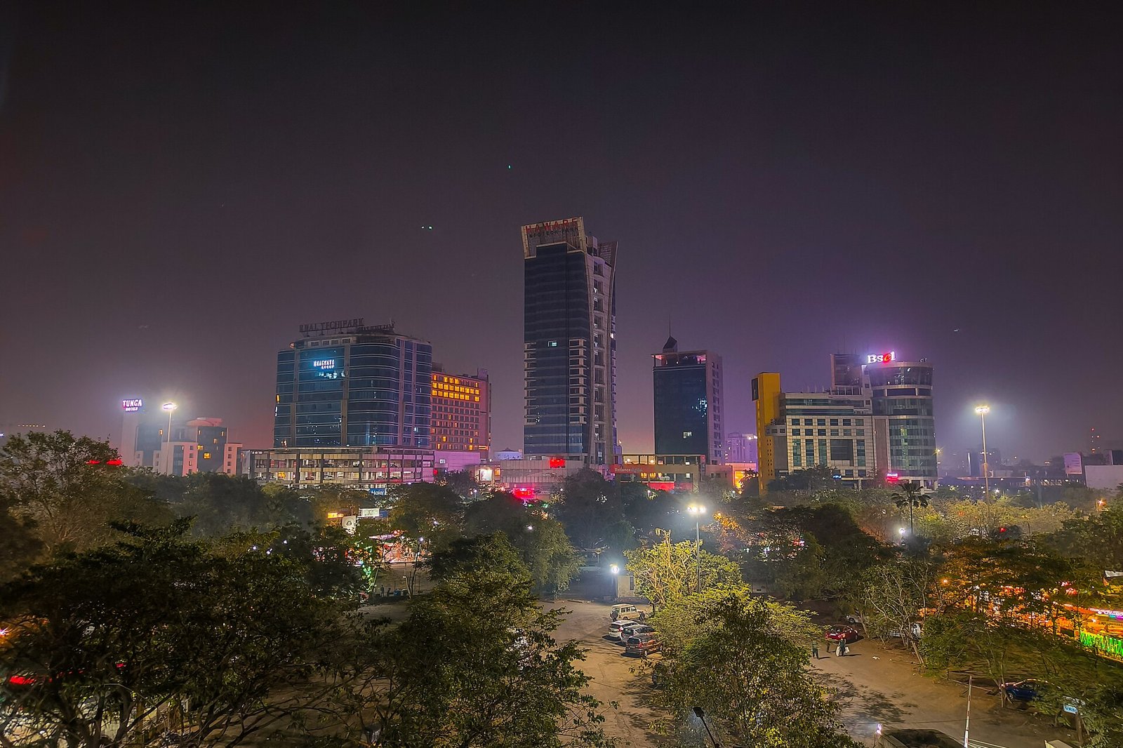 Tall buildings lit up at night in Vashi, seen over trees and a car park