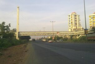 A cable stayed footbridge crossing a wide highway at Kharghar with apartment towers and trucks on the road