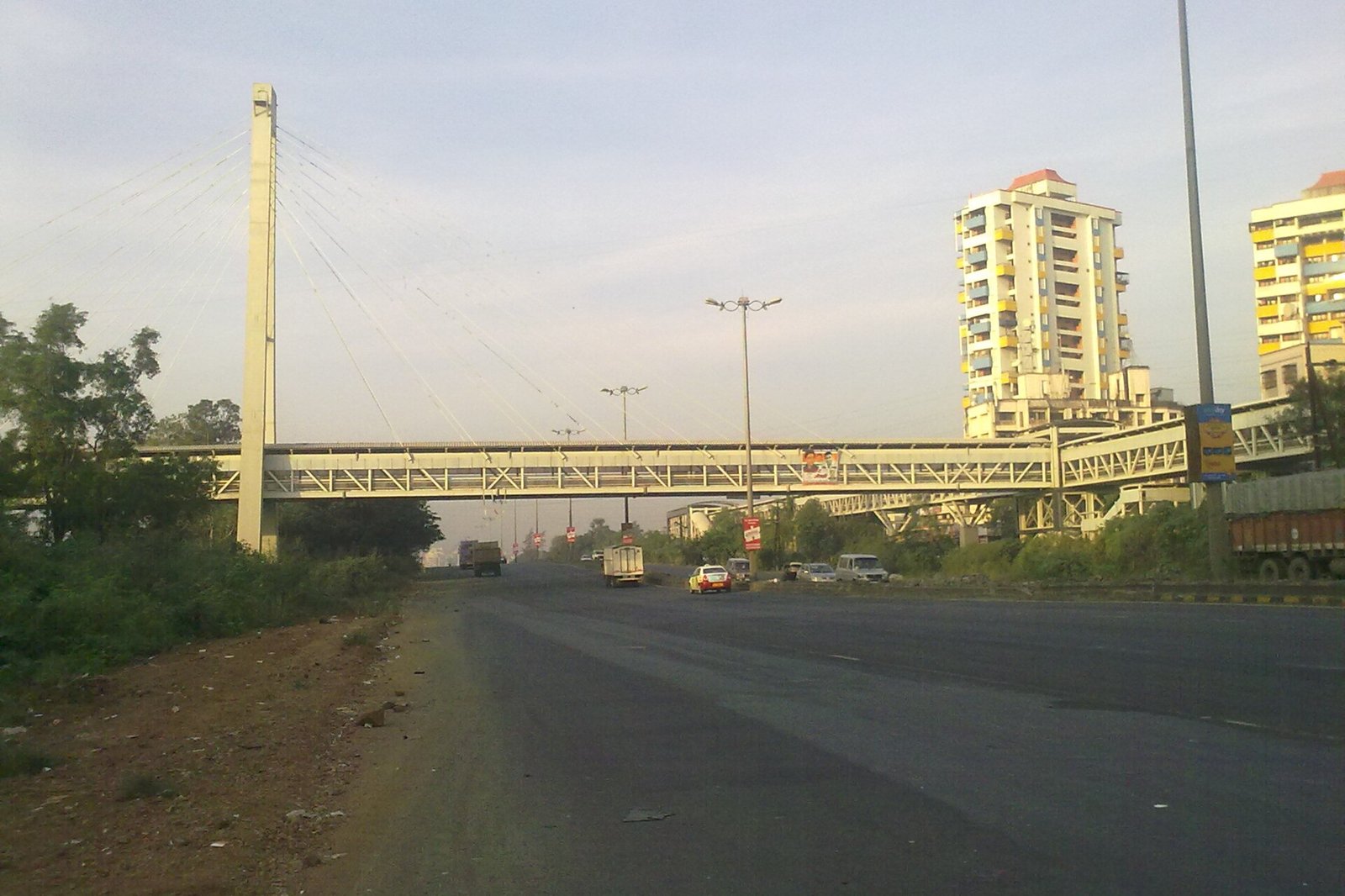 A cable stayed footbridge crossing a wide highway at Kharghar with apartment towers and trucks on the road