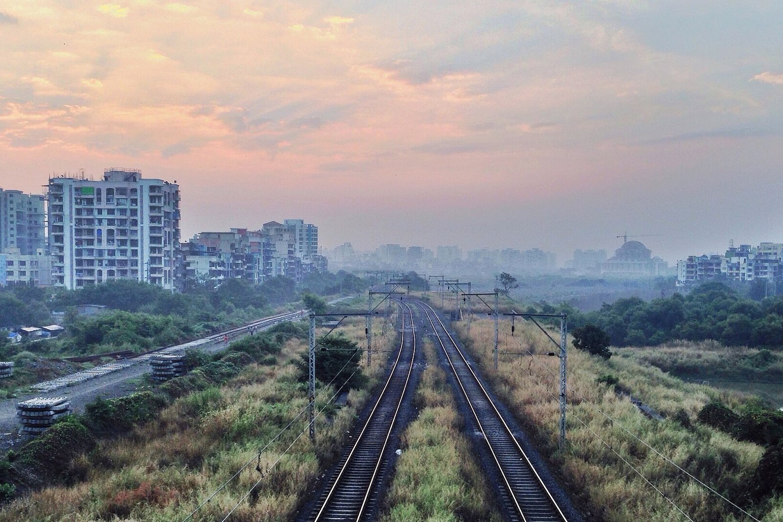 Railway tracks running past apartment towers and scrub under a hazy dawn sky near Seawoods in Navi Mumbai