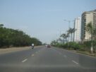 A wide multi lane road with a green median beside tall apartment buildings set back behind trees and palms on Palm Beach Road