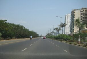 A wide multi lane road with a green median beside tall apartment buildings set back behind trees and palms on Palm Beach Road