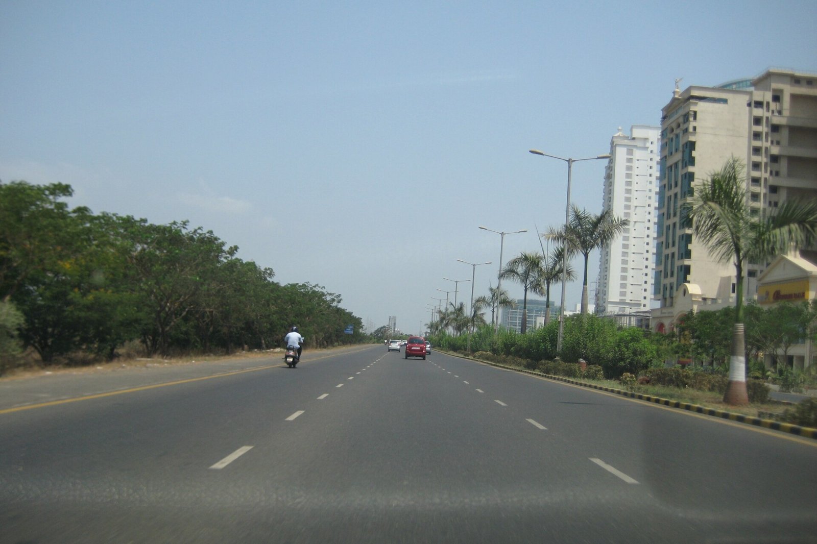 A wide multi lane road with a green median beside tall apartment buildings set back behind trees and palms on Palm Beach Road