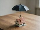 A small open umbrella held over a model house on a wooden table