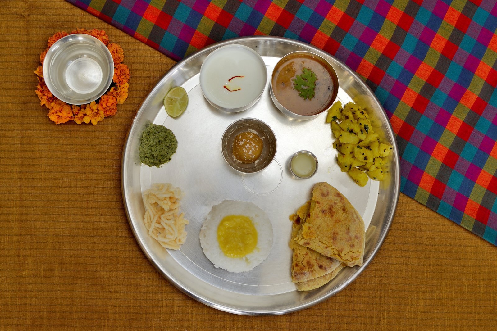Festive Maharashtrian meal on a steel thali with rice, sweets, flatbread, curry and chutneys, beside a bowl ringed with marigolds