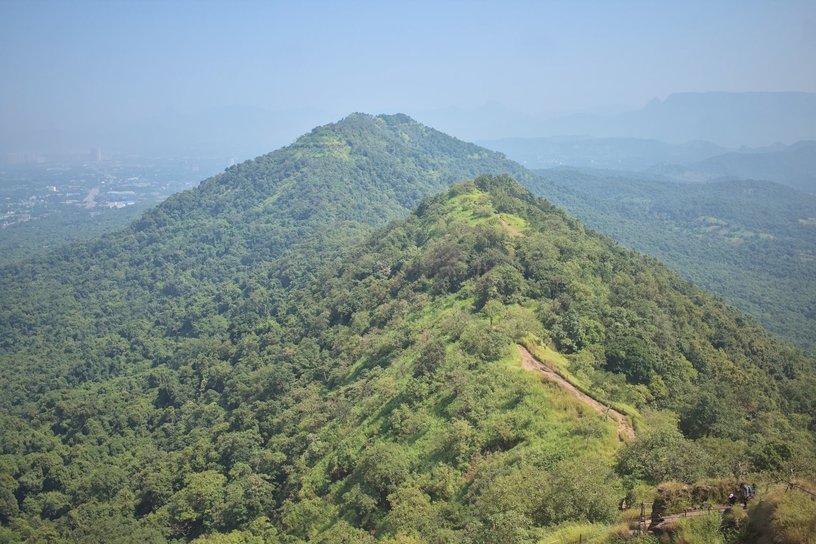 Forested ridge and a dirt trail seen from Karnala Fort, with hazy hills and the plain in the distance